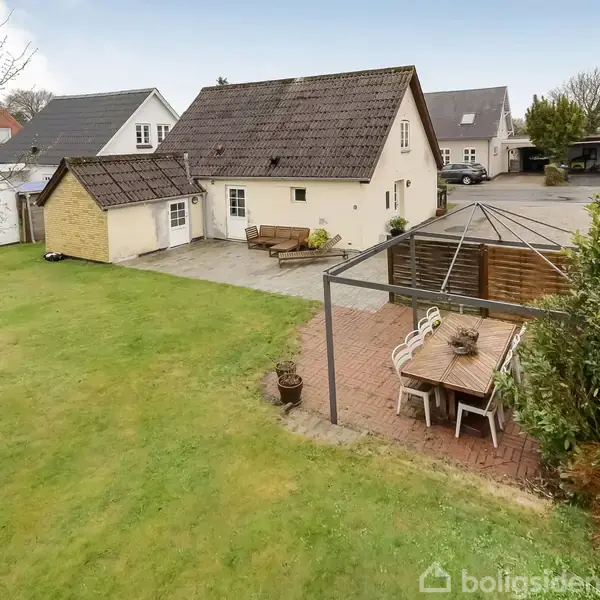 Hus med brun tag og gul facade står i en have. Et udendørs spisebord med stole er placeret under en pergola. Flere omkringliggende huse kan ses i baggrunden.