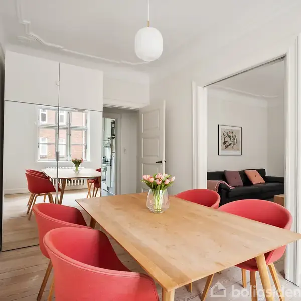 Et spisebord i træ med seks røde stole omkring det. En vase med blomster står på bordet. Rummet har hvide vægge, en stor spejlflade og en sofa i baggrunden.