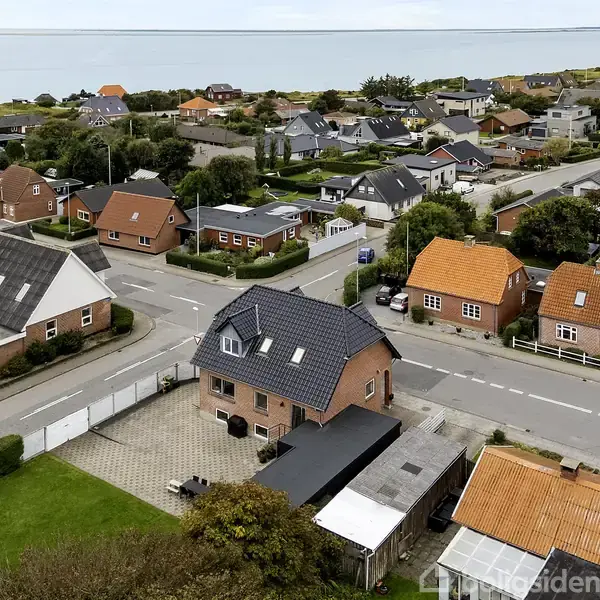 Huse ligger tæt samlet i boligkvarter, set fra luften. Et centralt hus med skråt sort tag har en gårdhave. Omkring er veje og grønne områder, og havet ses i horisonten.
