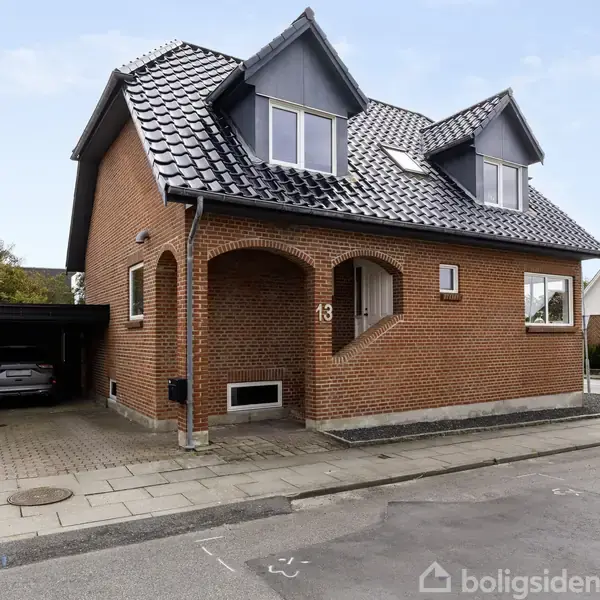 Rødt murstenshus med tre gavlvinduer, sorte tagsten, og nummer 13 ved indgangen. En bil holder i carport ved siden af. Huset ligger i et byområde med fortov og vej.