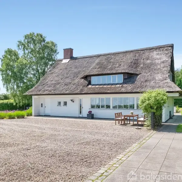 Et hus med stråtag, flankeret af grønne områder og træer, står ved en grusbelagt gårdsplads med en terrasse og bord-bænkesæt. Solrig dag.