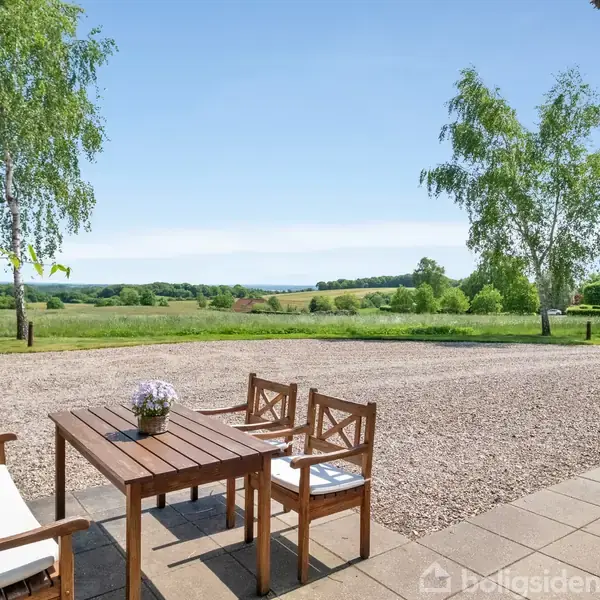 En træbord med stole står på en terrasse ved siden af en grusplads. Træer omkranser området, og landskabet strækker sig ud i en frodig, grøn horisont under en blå himmel.