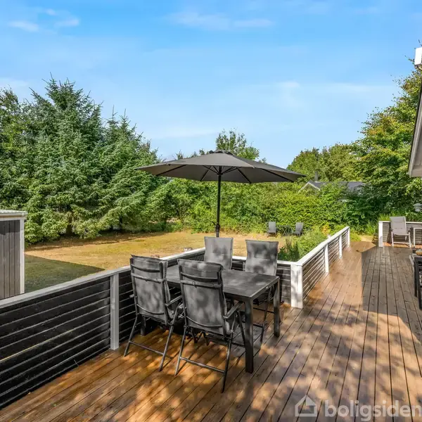Terrasse med sort træbord og seks stole under en sort parasol. Det er sommer, omgivet af grøn vegetation og op ad et gråt træhus. Skur til venstre.