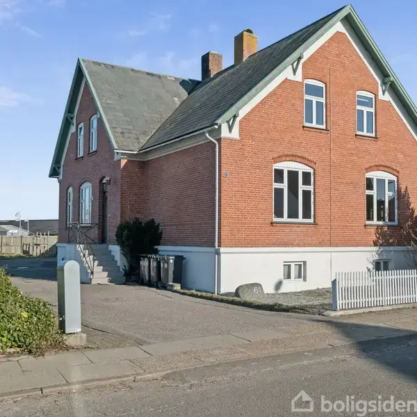 Et rødt murstenshus med skråtag og hvide vinduer står i sollys. Det har en hvid base og en lille baghave med en hvidt stakit.