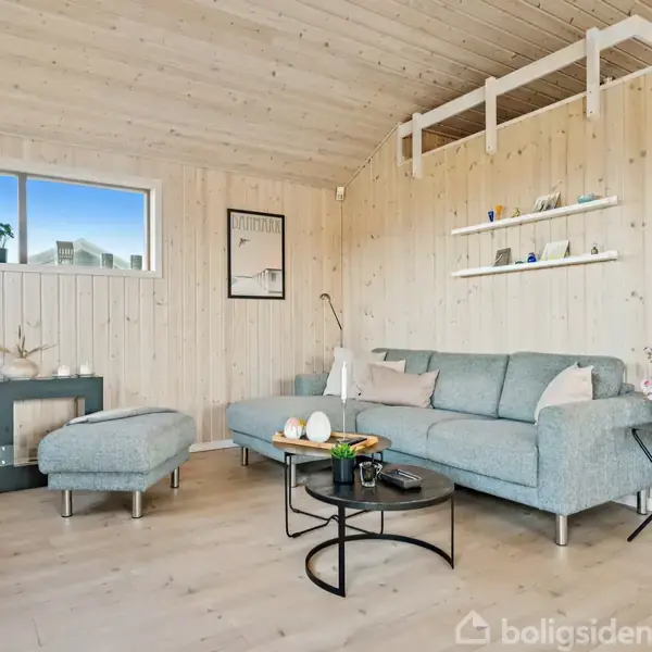 Sofagruppe med lyseblå sofa og skammel, placeret i en stue med lyse trævægge og loft. Rundt sofabord, vægmonterede hylder med dekorationer, og et stort vindue med udsigt udenfor.