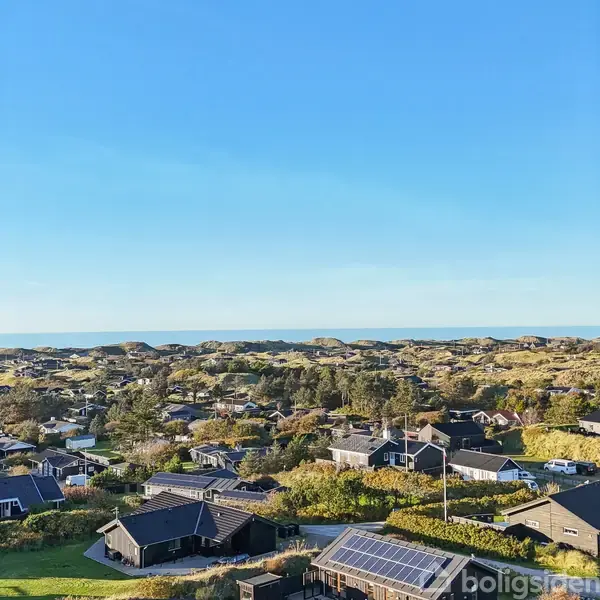 Landsbyhuse spredt mellem træer og buske; kystlinje og klitter i baggrunden under en klar blå himmel.