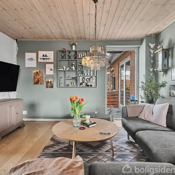 Gråt sofaarrangement står foran et rundt træbord med tulipaner, omgivet af bøger og dekorationer på en grøn væg. Fladskærms-TV på kommode. Lysekrone hænger ned fra træloft.