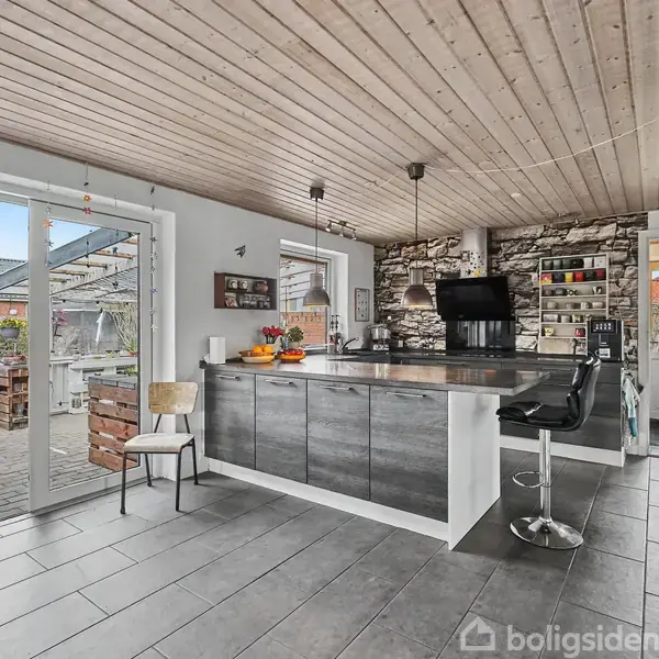 Køkken med moderne ø og barstol, beliggende i rum med stenvæg og lyse trælofter. Store glasdøre fører ud til en terrasse med udsigt over nabohuse.
