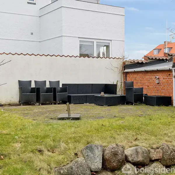 Havemøbler står arrangeret langs en hvid væg. En flisesti befinder sig i midten af en græsplæne, omgivet af sten og en rød murstensbygning ses i baggrunden.