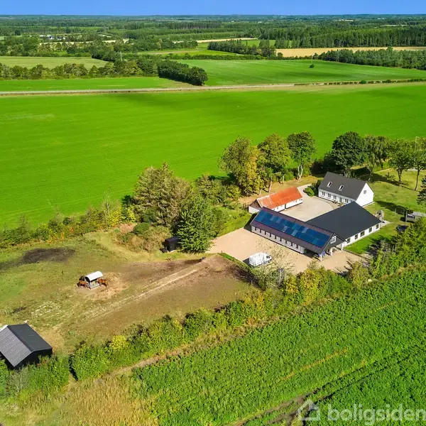 Luftfoto af en gård med flere bygninger omgivet af åbne grønne marker og træer. En indhegnet gårdsplads og mindre strukturer ligger tæt på hovedbygningerne.