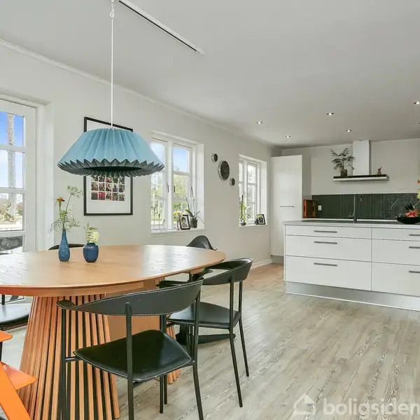 Et moderne køkken-alrum med et rundt træbord, omgivet af orange og sorte stole. Et blåt pendel hænger over bordet. Lyse skabe og trægulv danner rummet med store vinduer.