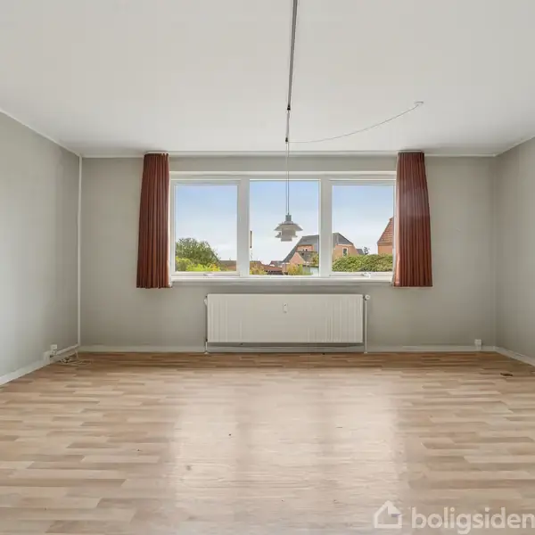 Stort, tomt rum med trægulv. Et vindue med røde gardiner ser ud mod hustage og himmel. En radiator er under vinduet, og der er en lampe hængende fra loftet.