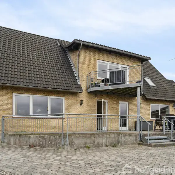 Toetagers murstenshus med valmtag, stor altan og rampe. Huset ligger på en flisebelagt grund med en trailer i forgrunden. Himlen er overskyet.