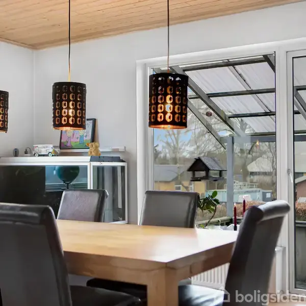 Et spisebord med sorte stole står i et rum med tre pendellamper hængende over. Gennem glasskydedøre ses en udendørs terrasse og haveområde.