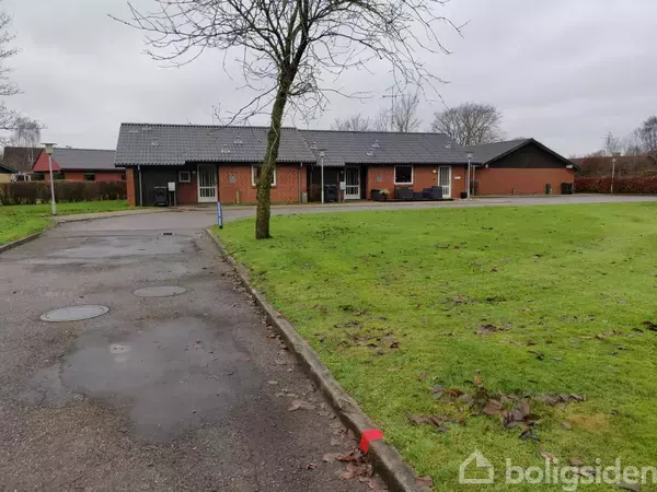 Rækkehus i røde mursten med sort tag ligger ved en grøn græsplæne. Træer uden blade ses i nærheden, og en asfalteret sti fører til bygningen.