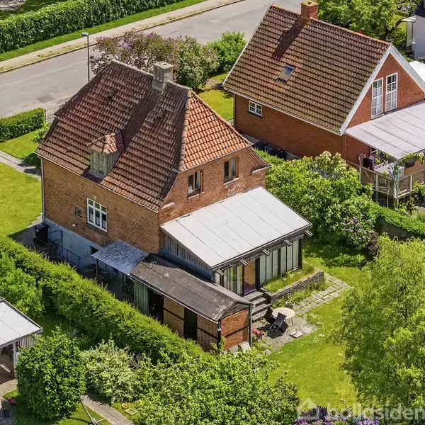 Rødt murstenshus med skråt tegltag står i grøn have. En vinterhave er vedhæftet. Omkring huset er velplejede buske og træer. I nærheden er andre lignende huse langs en vej.
