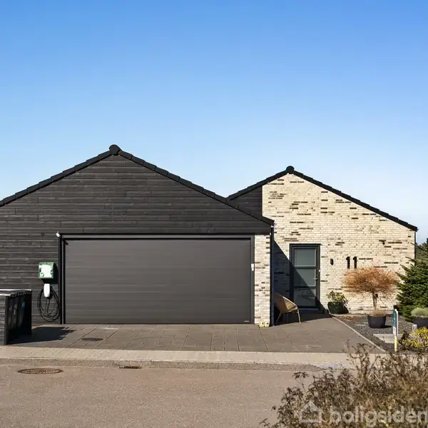 Et moderne hus med en dobbeltgarage i sort træ og stenfacade, står på en stille vej. Tal 11 vises på muren. Omkringliggende natur med blå himmel i baggrunden.