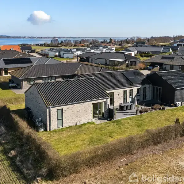En moderne, sort-tagget villa står roligt på en grøn græsplæne. Den er omringet af buske og andre huse i et beboelsesområde. I baggrunden ses en udsigt til vandet.