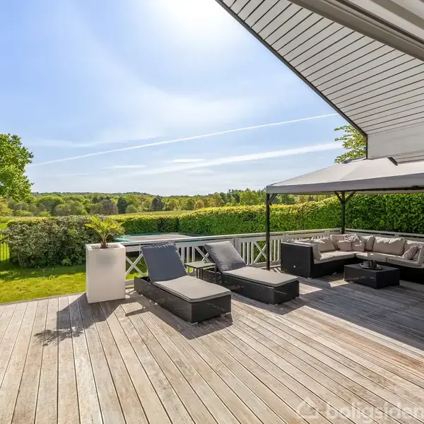 En terrasse med liggestole og en sofa under et pavillon-tag. Omkranset af grønne planter og en frodig græsplæne med udsigt til et åbent landskab.