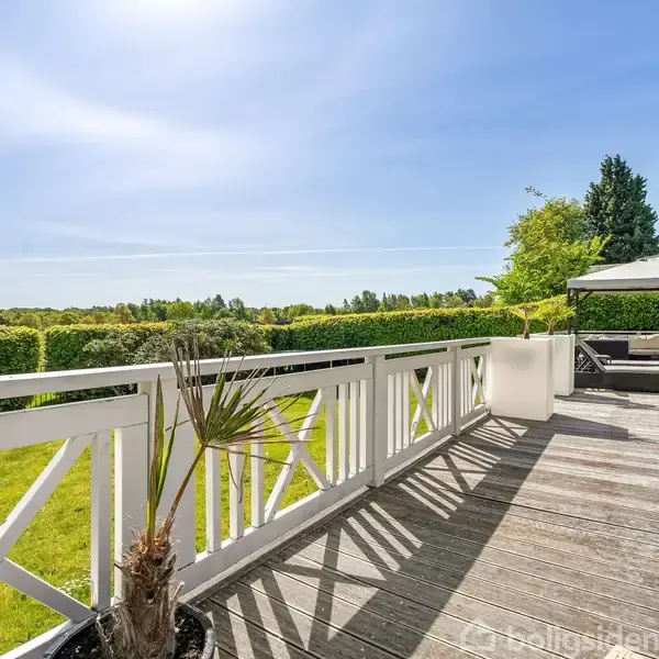 En stor træterrasse med hvide gelændere, omkranset af grønne buske. Terrassen har solsenge og parasoller, under en klar blå himmel, med udsigt over landskabet.
