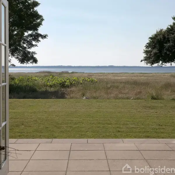 Åbne franske døre leder ud til en terrasse med fliser, der strækker sig over en græsplæne mod en bred udsigt over en sø eller hav og træer i baggrunden.