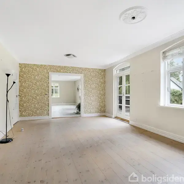 Stue med trægulv har en standardlampe ved siden af en væg med mønstret tapet. Der er store vinduer og en dør til en terrasse med udsigt over natur.