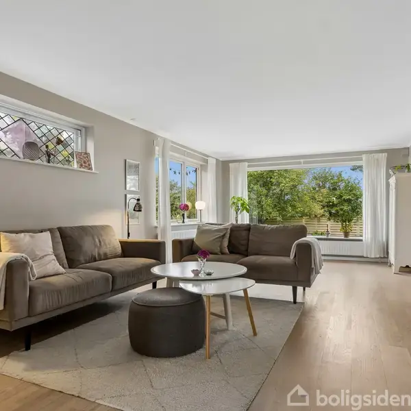 Stue med to brune sofaer placeret omkring et rundt sofabord, belyst af en standerlampe. Store vinduer giver udsigt til en grøn have.