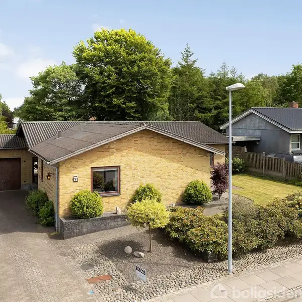 En gul murstenshus med sort tag står stille omgivet af en anlagt have og nabohuse. Grussti fører til indgang. Adresseplade viser "13". Tæt bevoksning bagved.