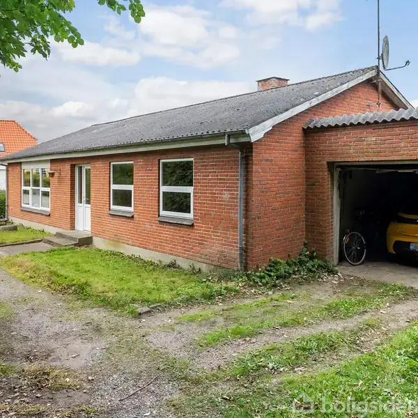 En rød murstensbygning med en garage, der indeholder en bil og en cykel. Bygningen har flere vinduer og en hvid dør. Omgivelserne er en have med græsplæne og indkørsel.