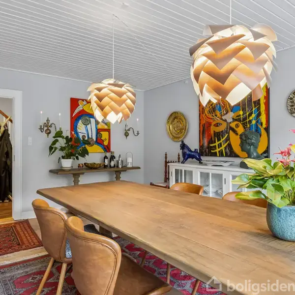 Et spisebord i træ er omgivet af fem brune stole, stående under to store pendellamper. Væggene har kunstværker, og planter dekorerer rummet, der har et lyst, nordisk design.