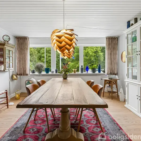Træbord med vase står i spisestue med farverig tæppe og designerlampe. Omringet af glasvitriner, stole, kunstgenstande og store vinduer med udsigt til grøn have.