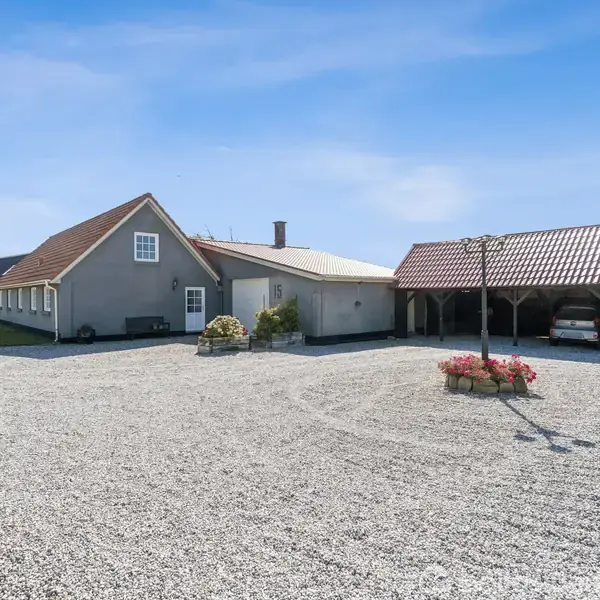 Et gråmalet hus med rødt tag står stille ved siden af en carport med en bil. Gruset gårdsplads omgiver bygningerne, mens blomsterbedet pryder midten.