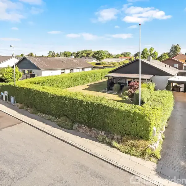 En villa med fladt tag er omgivet af en velplejet hæk. Foran huset er en indkørsel og en garage på den højre side. Området er et forstadskvarter med flere lignende huse.