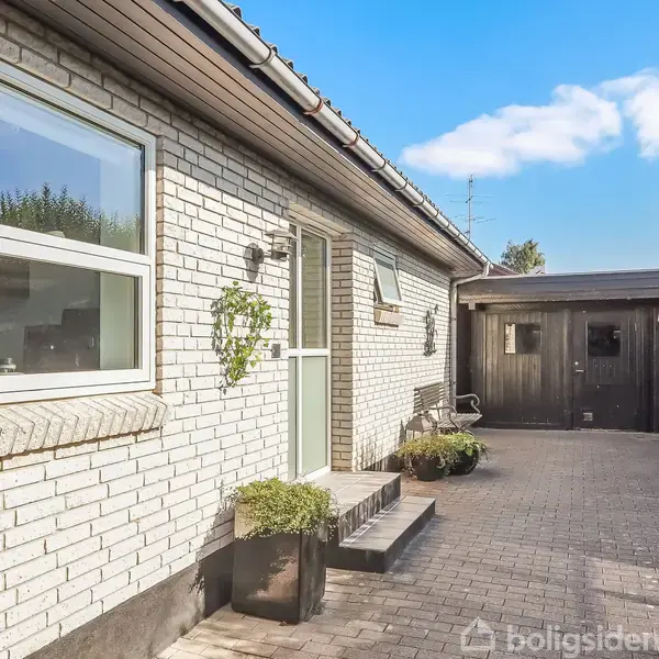 Hus med hvid murstensfacade har en grøn dør og vinduer, omgivet af en flisebelagt indkørsel. I baggrunden ses en trægarage og grønne buske under en blå himmel.