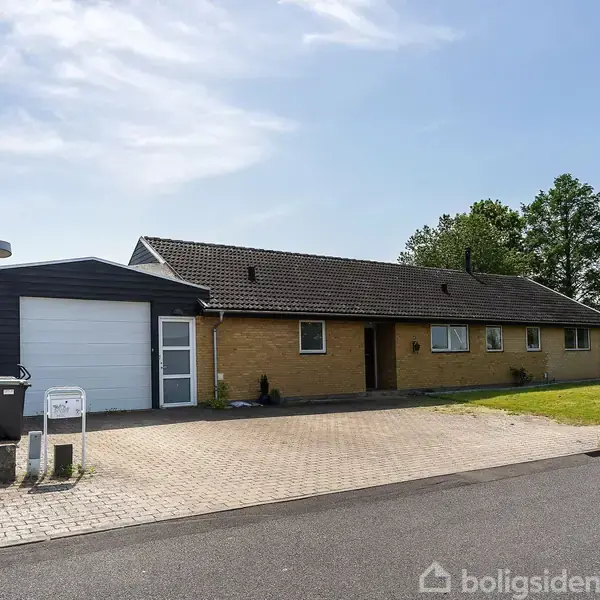 Et gulstenshus med garage står langs en vej. Det har et sort tag og en flisebelagt indkørsel. Omgivelserne inkluderer en græsplæne og træer i baggrunden.