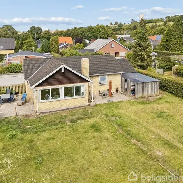 Et ældre parcelhus med gavl udført i træ, omgivet af en have og hække. Huset ligger i et villakvarter med flere lignende boliger og grønne områder i baggrunden.