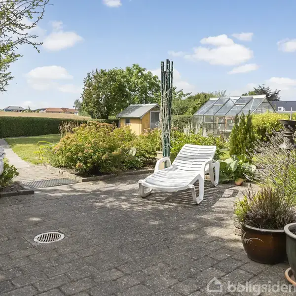 En hvid liggestol står på en flisebelagt terrasse. Omkring den er en have med planter, en glasdrivhus og en havefigur af en hane. Det er solrigt og frodigt.