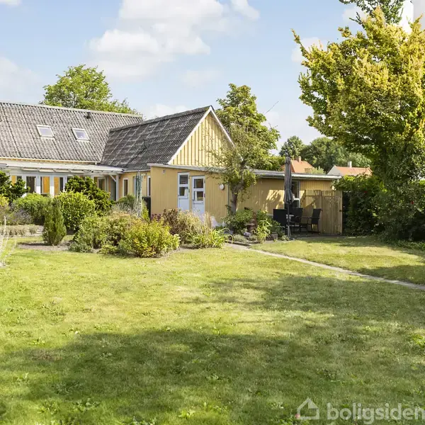 Hus med gul facade og mørkt tag står i en frodig have med græs og buske. Havemøbler og parasol er placeret ved siden af huset under en klar himmel.