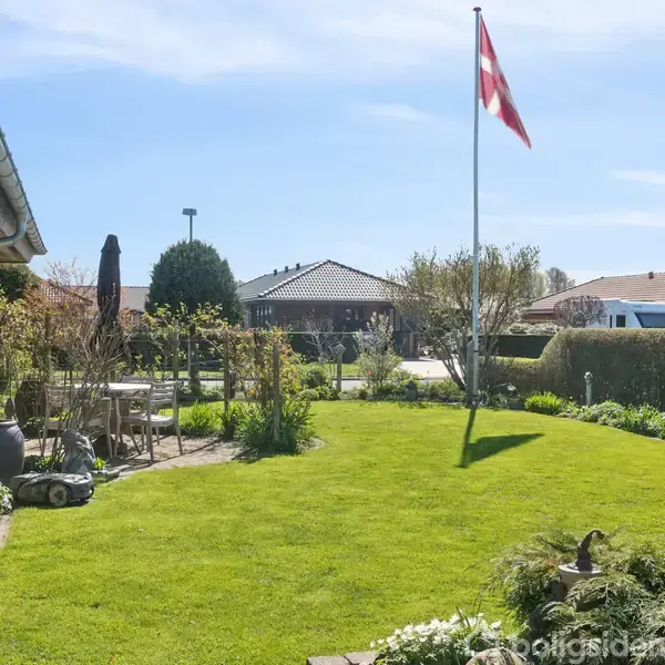 En flagstang står med Dannebrog vajende i en velplejet have. Til venstre løber et stisystem ved huset, og i baggrunden ses flere huse og en parkeret bil under en blå himmel.