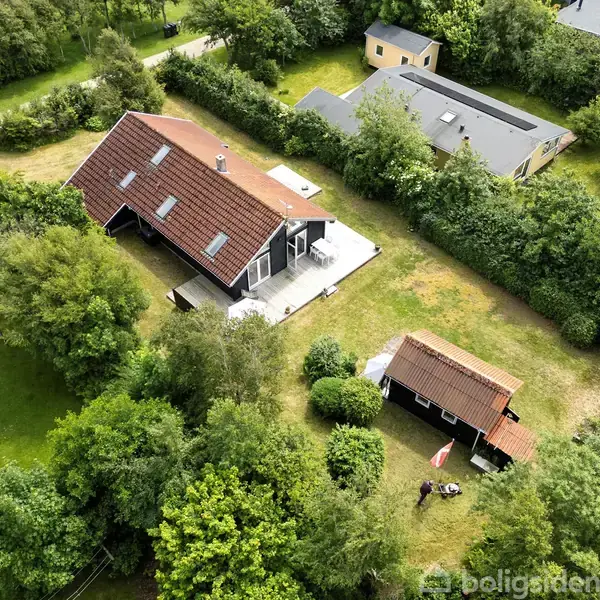 Hus med rødt tag omgivet af frodig have og grønne træer, placeret i en række af lignende bygninger i et skovklædt område. En lille terrasse ses ved siden af huset.