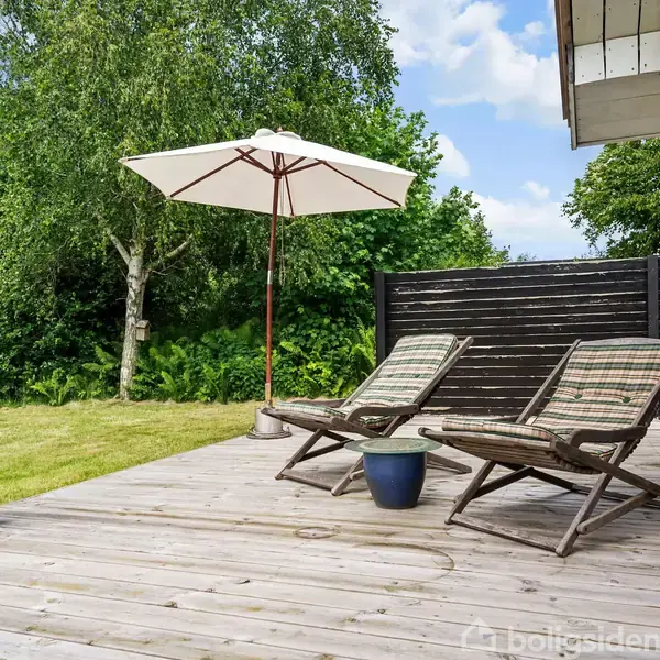 To liggestole står på en træterrasse med en parasol. Omkring er der en grøn have med træer og buske. Et lille bord med en plante står mellem liggestolene.