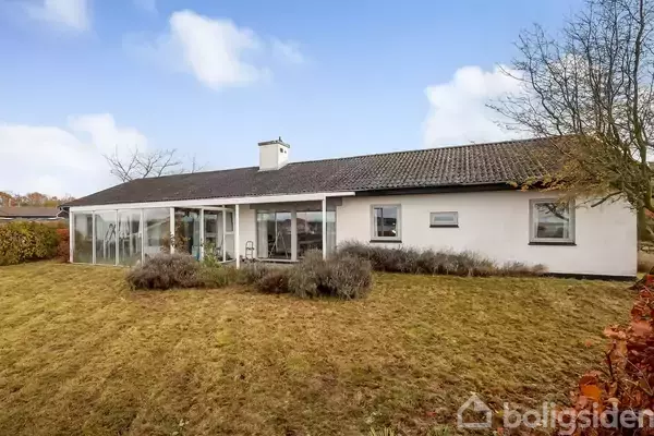 Et hvidt en-etagers hus står stille i en have med græsplæne og buske. Huset har store vinduer langs facaden, omgivet af træer.