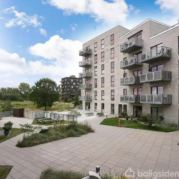 Lejlighedskomplekser står roligt omkring en fælles gårdhave med flisebelægning og grønne områder. Cykler står parkeret op ad bygningerne under en blå, skyet himmel.