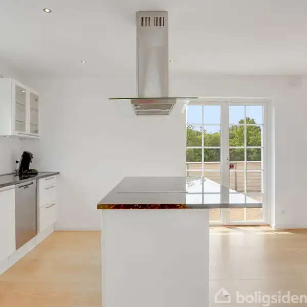 Et moderne køkken med en central kogeø og emhætte. Store vinduer bagtil giver udsigt til træer og en terrasse. Lyse trægulve og hvide skabe skaber et rent udtryk.