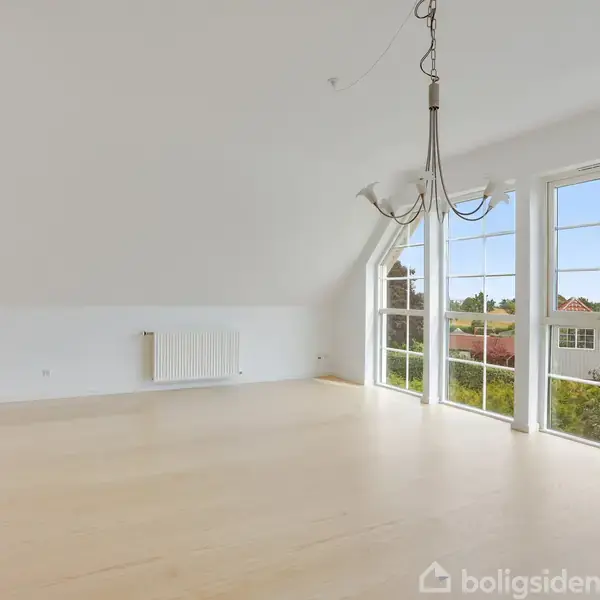 Et tomt rum med en ophængt lysekrone, masser af naturligt lys strømmer ind gennem store gulv-til-loft-vinduer. Udenfor ses tage og træer.