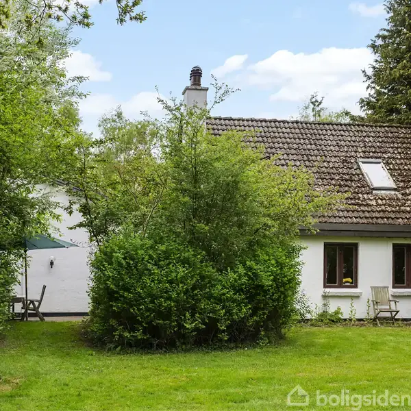 Et hvidt hus med brun teglstenstag står omgivet af frodig have og høje træer, mens havemøbler er placeret på græsplænen foran huset.