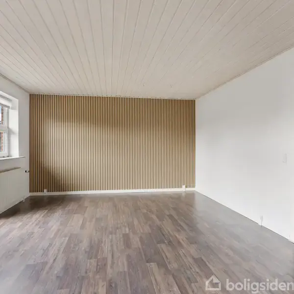 Stue med trægulv og loft, lyst fra store vinduer til venstre. Væg med lodrette træpaneler. Åbning fører til køkken i højre hjørne.