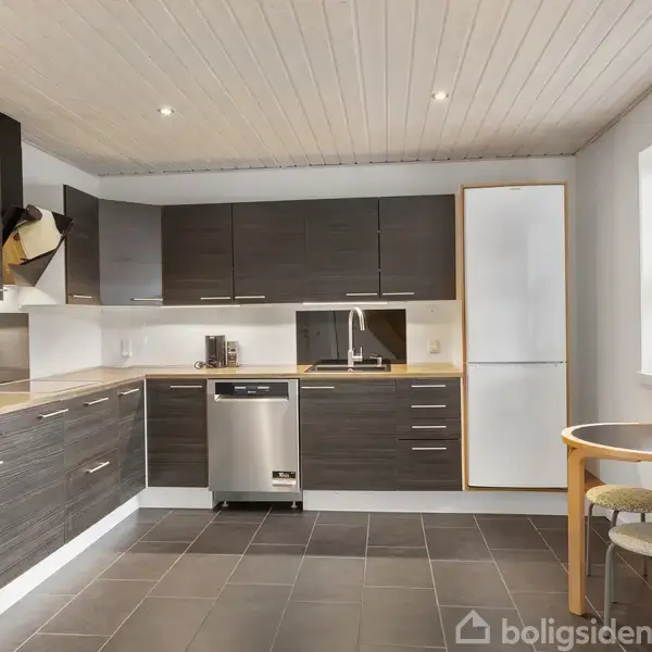 Køkken med mørke træskabe installeres langs to vægge; en ovn og opvaskemaskine er indbygget. I baggrunden er et stort vindue. To taburetter står ved et lille rundt bord.