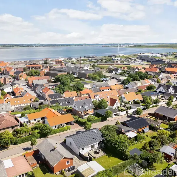 Boligområde med røde og grå hustage, set fra oven, ved siden af en kystlinje med vand i baggrunden. Området er omgivet af grønt og træer under en blå himmel med skyer.