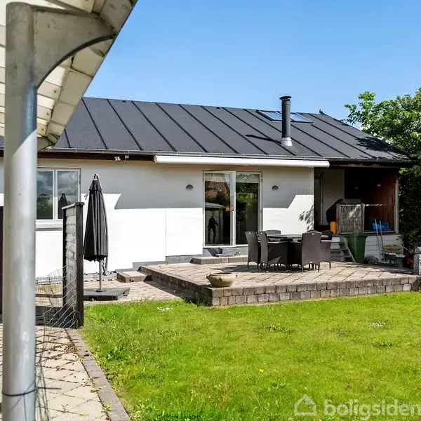 Huset står med hvid facade og sort tag. Foran er en terrasse med havemøbler, omgivet af grøn græsplæne. Til venstre ses en flisebelagt gangsti og en carport.